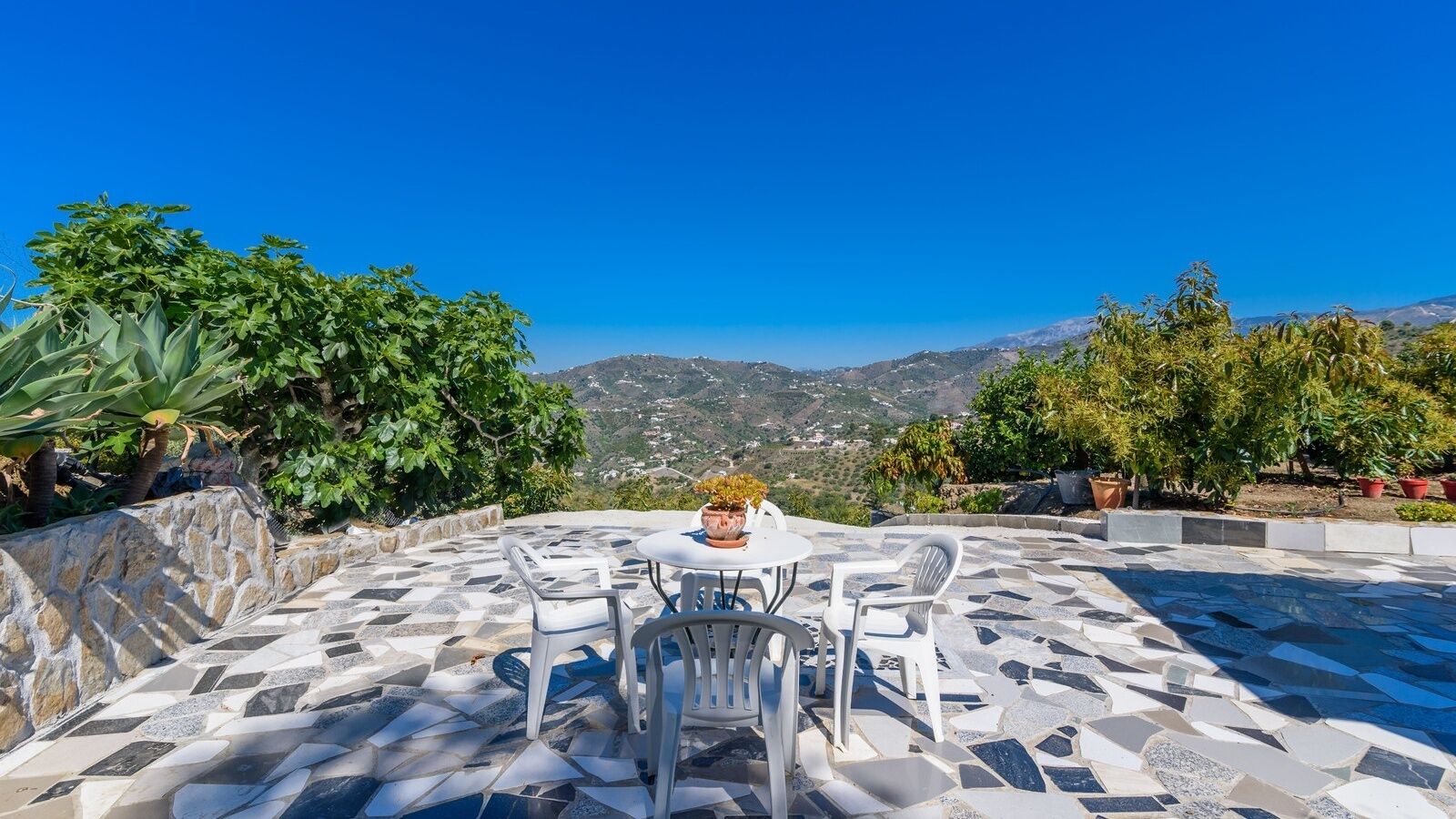 A terrace with a stone floor, white chairs, and a table with a potted plant.