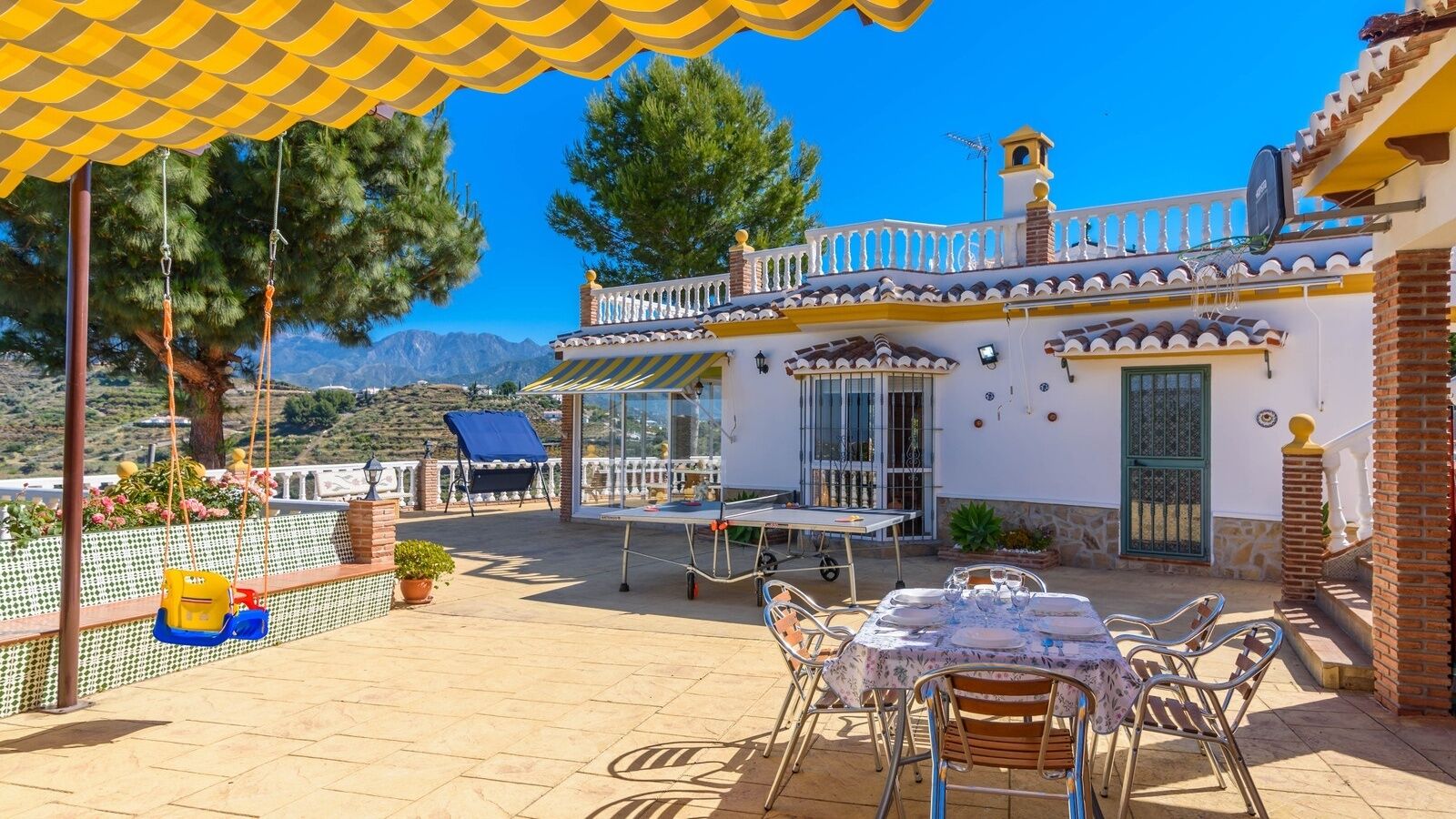A patio with a table set for dining, a swing set, and a view of the mountains.