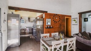A modern kitchen with stainless steel appliances, a brick accent wall, and a dining area with white chairs and a wooden table.