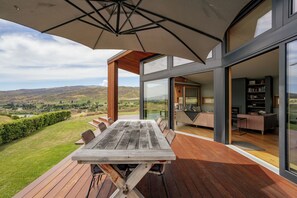 A modern outdoor dining area with a wooden table and chairs, a large umbrella, and a scenic view of the countryside. - Vino and Views  - Bannockburn Holiday Home (Cromwell)