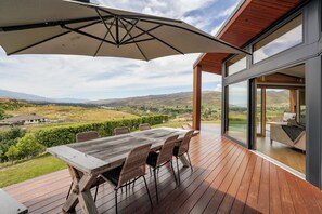 A wooden deck with a table and chairs, a large umbrella, and a view of a valley and mountains. - Vino and Views  - Bannockburn Holiday Home (Cromwell)