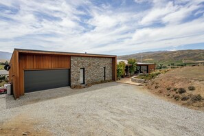 A modern house with a large garage, stone exterior, and a gravel driveway. - Vino and Views  - Bannockburn Holiday Home (Cromwell)