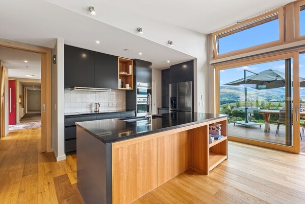 A modern kitchen with a large island, dark cabinetry, and a view of the outdoors through large windows.