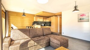 Una sala de estar moderna con un sofá modular, vigas de madera en el techo, una zona de cocina e iluminación colgante.