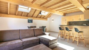Un moderno apartamento tipo loft con techo de madera, un sofá modular, una zona de comedor con taburetes de bar y una cocina con armarios de madera.