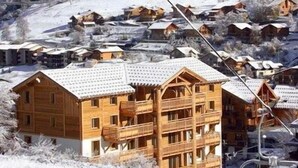 Un chalet in legno con un balcone, circondato da case innevate e una seggiovia sullo sfondo.