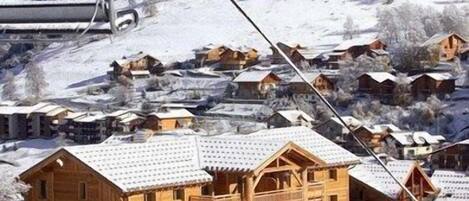 Un chalet en bois doté d’un balcon, entouré de maisons recouvertes de neige, avec un télésiège visible au loin.
