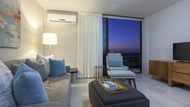 A modern living room with a sofa, armchair, and ottoman, a television, and a view of the evening sky through a large window. - Apartment with GREAT LOCATION, 1 block from the boardwalk (La Paz)