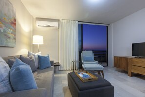 Ein modernes Wohnzimmer mit Sofa, Sesseln, Hocker, Fernseher und Blick auf den Abendhimmel durch ein großes Fenster.