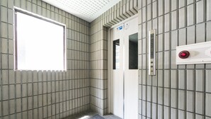 A narrow elevator with a tiled wall, a door with a red emergency button, and a small window allowing natural light. - Sapporo East Stay minpaku (Sapporo)