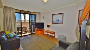 Sala de estar com sofá, televisão, mesinha e vista através de uma porta de vidro.