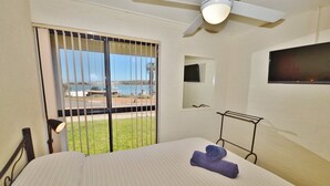 Quarto de hotel com cama, ventoinha no teto, televisão fixada na parede e vista para o exterior através de uma janela com grades.