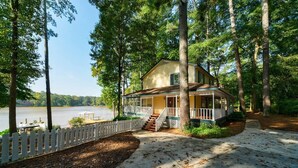 A lakeside cabin with a porch, surrounded by tall trees and a wooden fence. - LAY Back in the Lake House (Lilburn)