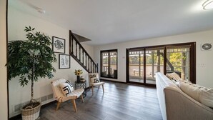 A spacious living room with a staircase, a large potted plant, a sofa, and a dining area visible through glass doors. - LAY Back in the Lake House (Lilburn)