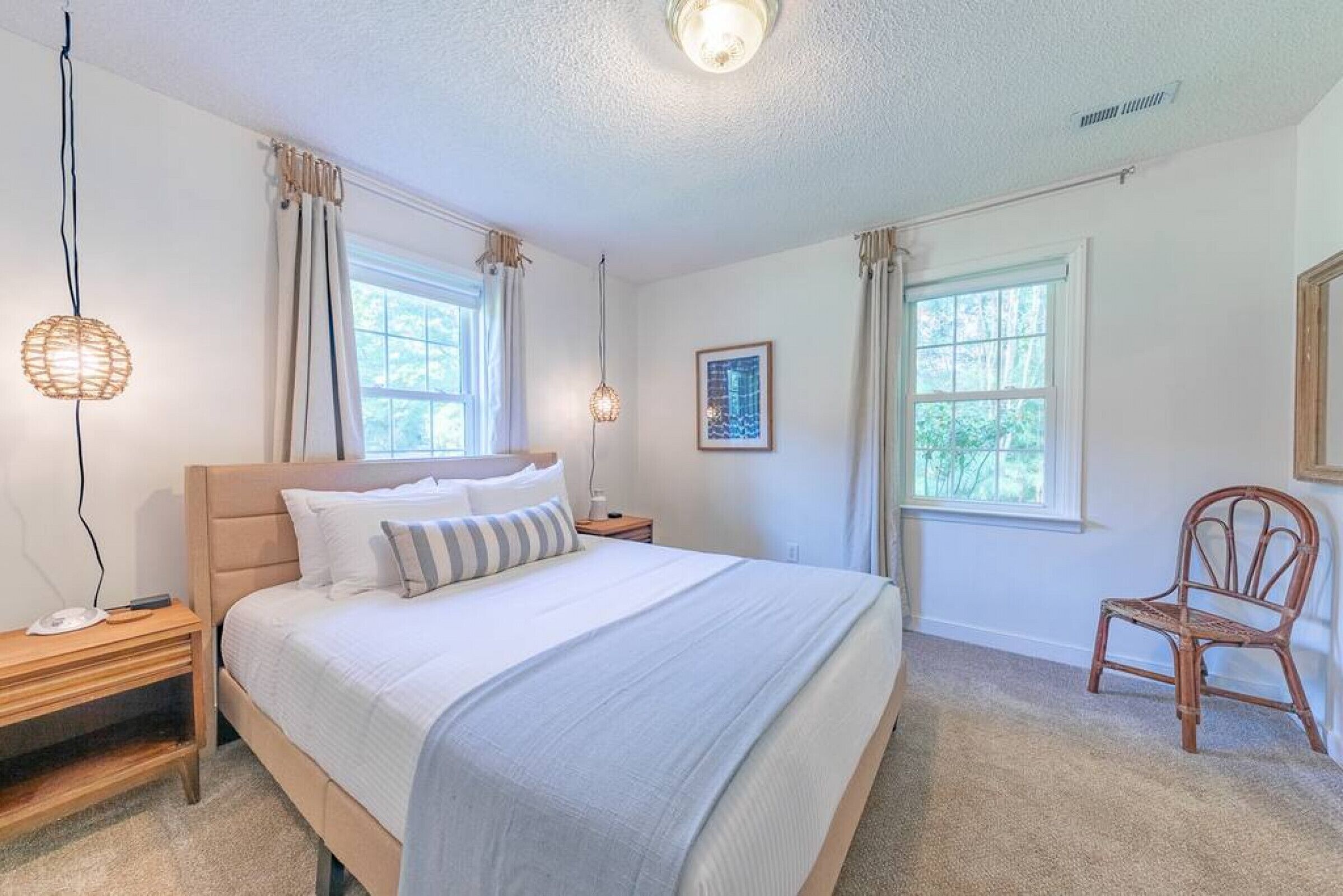 A bedroom with a large bed, two bedside tables, a chair, and two windows.