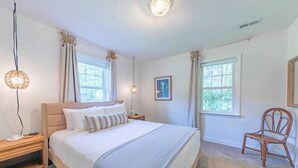 Ein Schlafzimmer mit einem großen Bett, zwei Nachttischen, einem Sessel und zwei Fenstern.