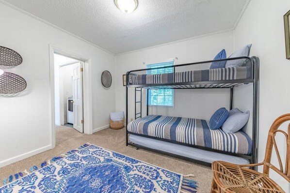 Habitación con literas, un tapete estampado, una silla de mimbre y una puerta que da a otra habitación.