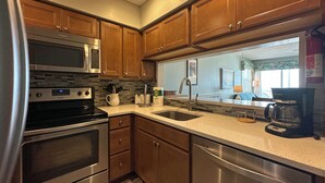 A modern kitchen with stainless steel appliances, wooden cabinets, and a large window providing natural light. - Captain's Walk I B1 - Oceanfront in Garden City Beach, SC (Garden City)