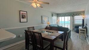 A dining area with a table and chairs, a ceiling fan, and a view of the ocean through a window. - Captain's Walk I B1 - Oceanfront in Garden City Beach, SC (Garden City)