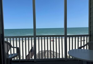 Vista a la playa desde un balcón cubierto con barandillas blancas, una pequeña mesa y dos sillas.
