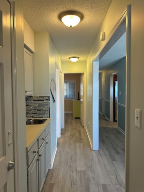 Ein enger Flur mit einer Küche links, einem Badezimmer mit Toilette und Waschbecken, und einem Schlafzimmer mit Bett und Kommode rechts.