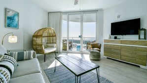 A spacious living room with a sofa, a coffee table, a wicker chair, a flat-screen TV, and a large window offering a view of the outdoors. - Boho Coastal Condo w/ Free Beach Shuttle at The Palms of Destin! 🐬🐚☀ (Destin)