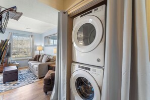 A laundry room with stacked washing machines, a sofa, a lamp, and a window with blinds.