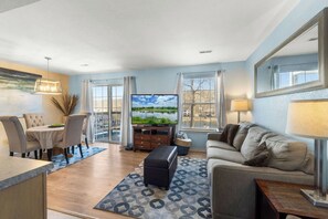 A modern living room with a flat-screen TV, a sofa, a dining area with chairs, and a large window with curtains.