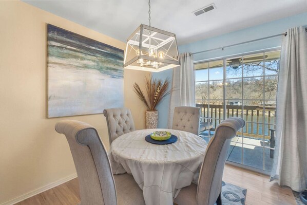 A dining area with a round table, four chairs, a hanging light fixture, a painting on the wall, and a view through a large window.