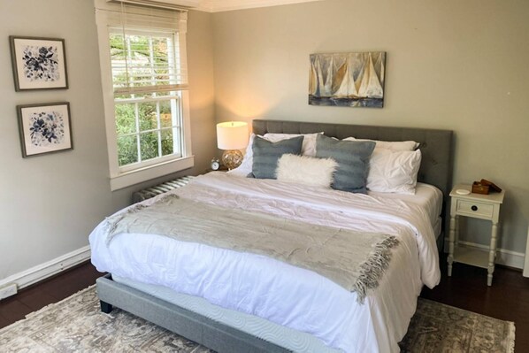 A bedroom with a large bed, a window with blinds, a bedside table with a lamp, and artwork on the wall.