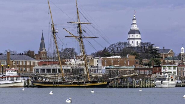Un'imbarcazione a vela storica ormeggiata in un porto, con un edificio bianco con cupola ben visibile sullo sfondo.