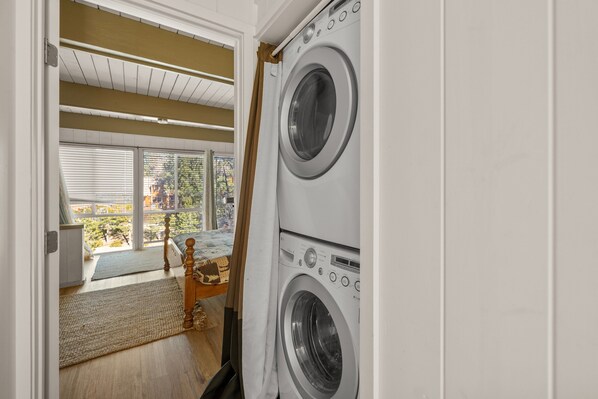 A laundry room with two stacked front-loading washing machines, a wooden bench, and a view of the outside through large windows.