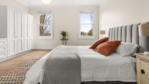Ein Schlafzimmer mit einem großen Bett, zwei Fenstern, einem Nachttisch mit Lampe und einer Topfpflanze.
