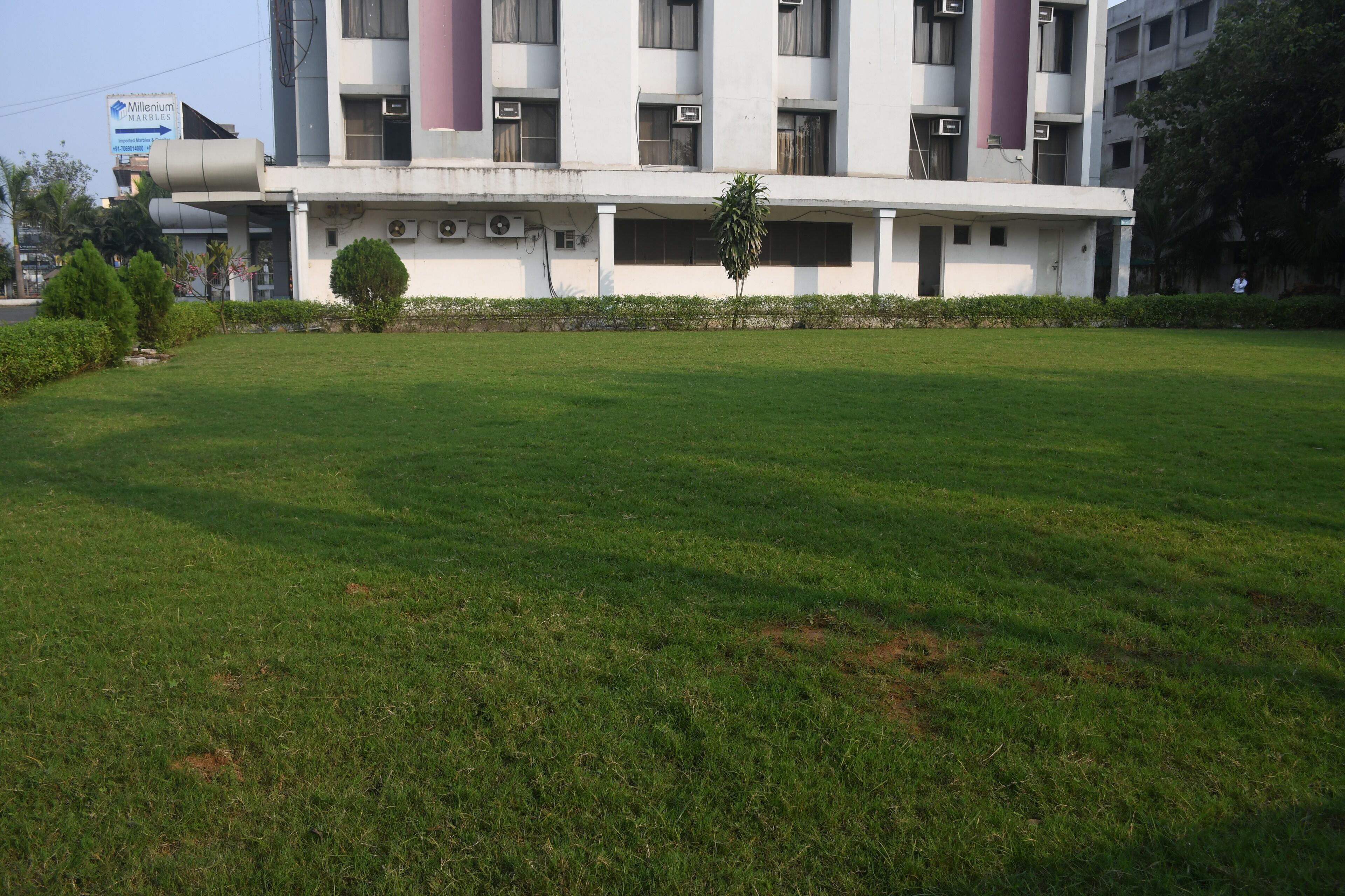 A building with a flat roof, multiple windows, and air conditioning units. There is a grassy area in front with a few small trees and a signboard in the background.