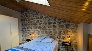 A bedroom with a stone wall, wooden ceiling, a bed with a patterned bedspread, two bedside tables with lamps, and a wardrobe.