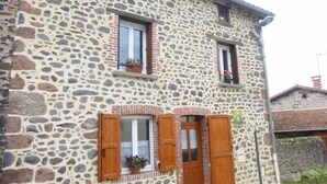 Un bâtiment en pierre doté de deux fenêtres, chacune ornée de volets en bois, et d’un petit balcon où est posée une plante en pot.