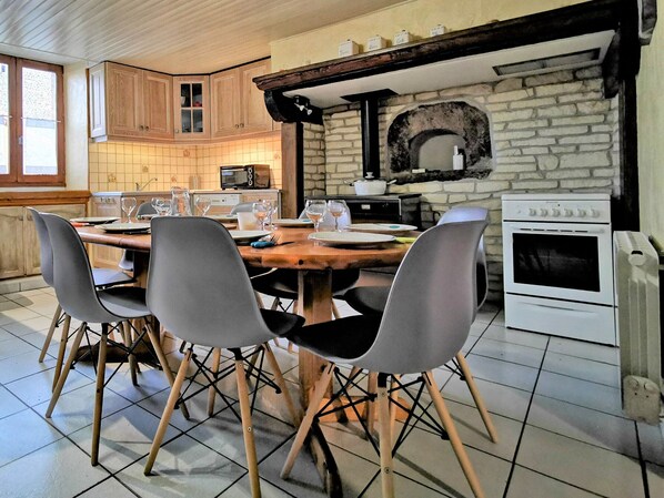 A dining area with a wooden table, four chairs, a kitchen with a stone wall, a microwave, a stove, and a window.