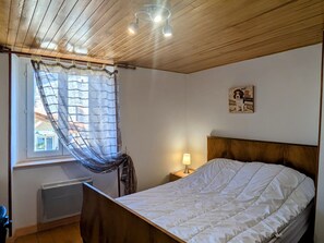 A bedroom with a wooden ceiling, a bed with a white duvet, a window with a sheer curtain, a wall-mounted picture, and a bedside lamp.
