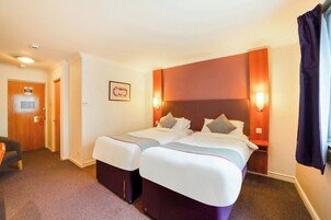 Habitación de hotel con dos camas, una puerta de madera, un cuadro en la pared y una mesita de noche con una lámpara.
