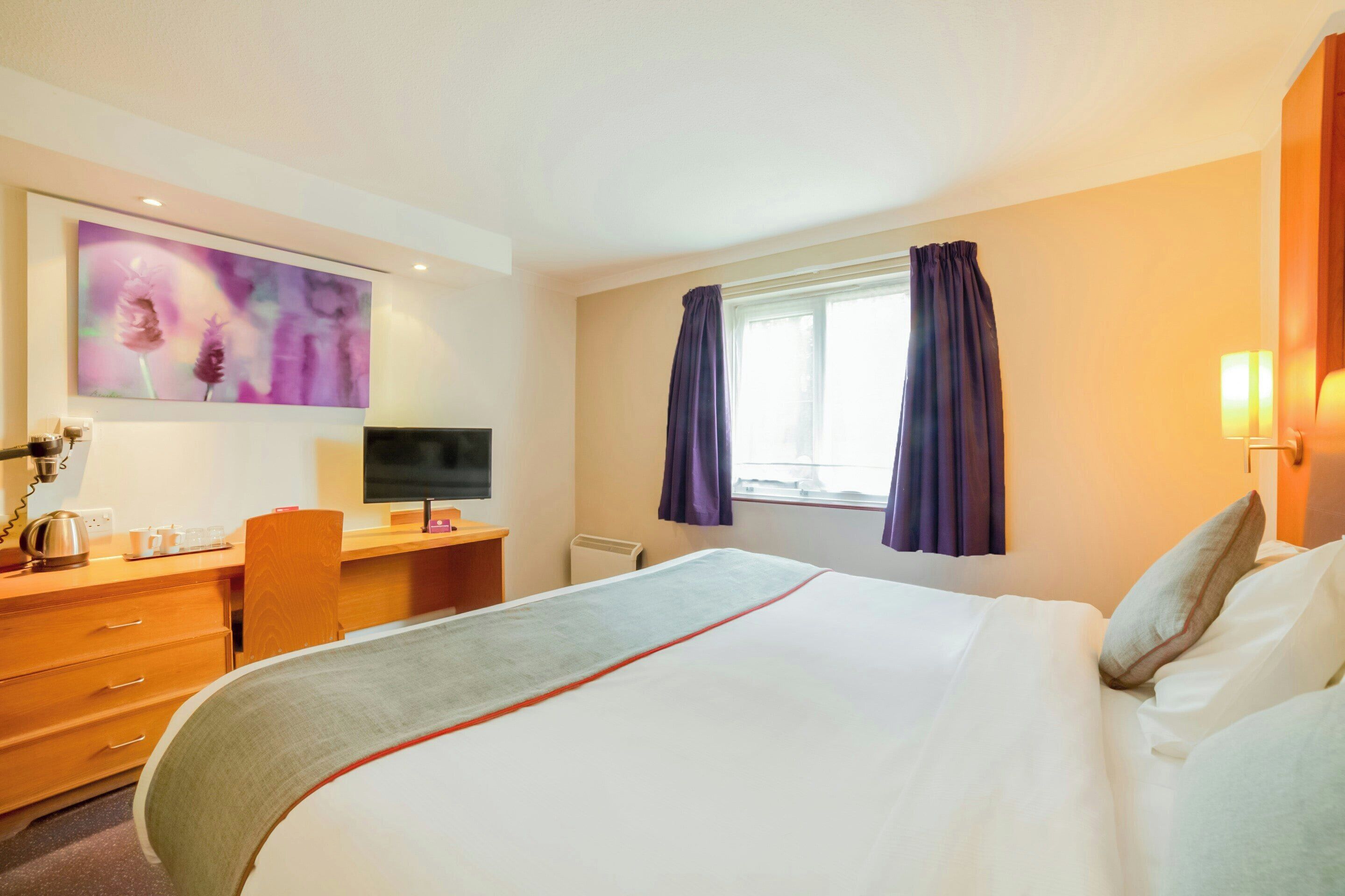 A hotel room with a bed, desk, chair, television, and artwork on the wall.