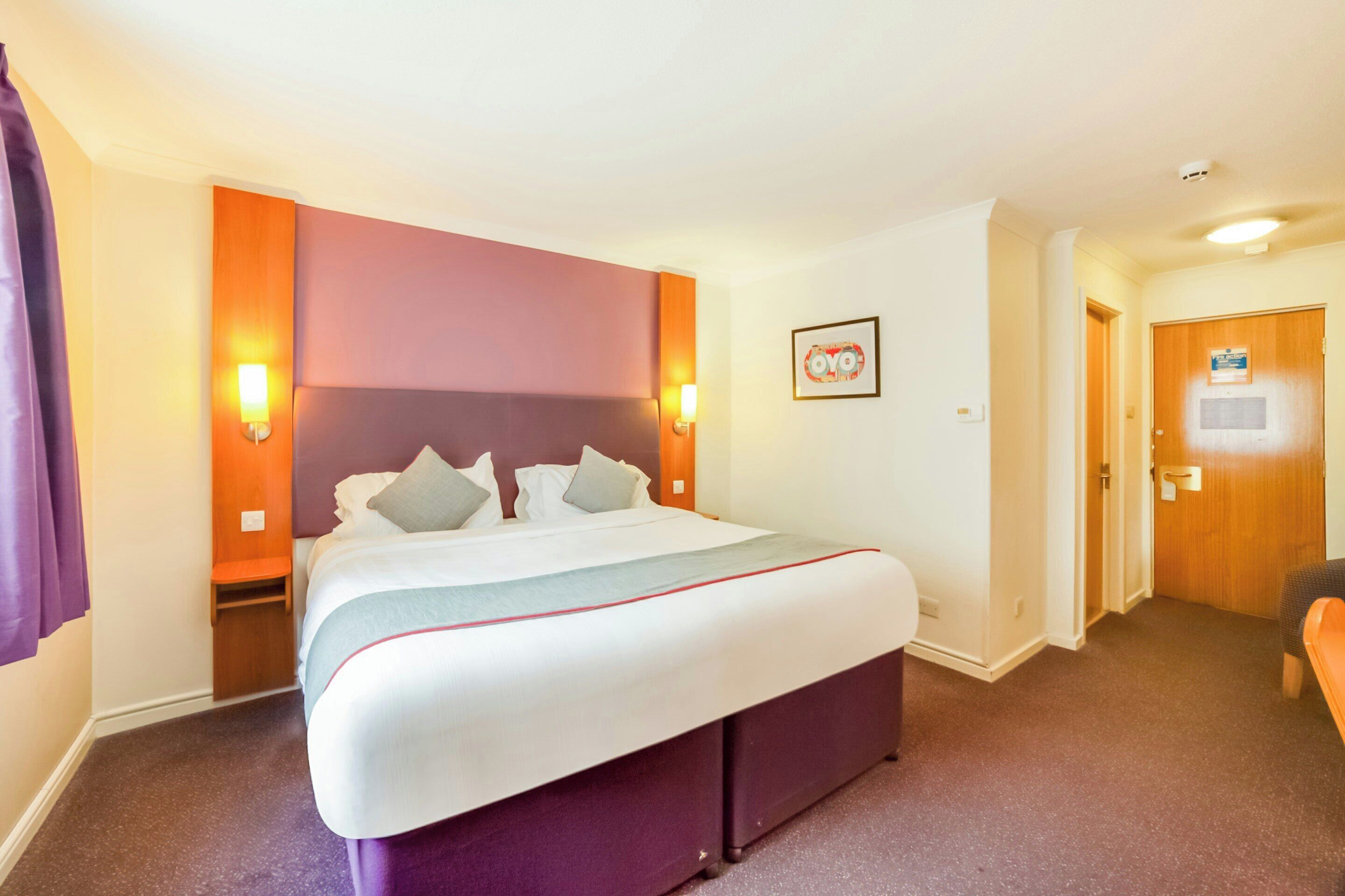 Quarto de hotel com uma cama grande, abajures, cabeceira, porta e uma pequena mesa.