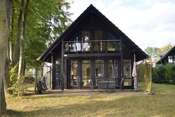 Un bâtiment en bois à deux étages, doté de grandes fenêtres, d’un balcon et d’un espace extérieur clôturé aménagé avec des chaises.