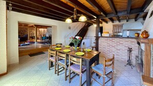 Ein Essbereich mit Holztisch, gedeckt für vier Personen, umgeben von Stühlen mit karierten Kissen. Rechts ist ein Kamin aus Backsteinen zu sehen, und im Hintergrund befindet sich eine Küche mit Backsteinwand und Holzschränken.