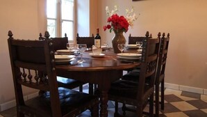 Une salle à manger avec une table ronde dressée pour six personnes, entourée de chaises en bois ornées de sculptures complexes. Un vase contenant des fleurs rouges et blanches est posé sur la table, et une bouteille de vin est visible. La pièce a un sol à carreaux et un tableau est accroché au mur.