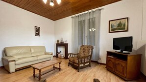 Un salon avec un plafond en bois, un canapé beige, un fauteuil à motifs floraux, une table basse en bois, un téléviseur posé sur un meuble en bois et une cheminée agrémentée d’objets décoratifs.