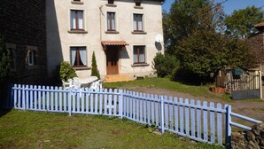 Une maison à deux étages avec une clôture en bois peinte en blanc, un petit patio avec des meubles d’extérieur et un mur de pierre sur le côté gauche.