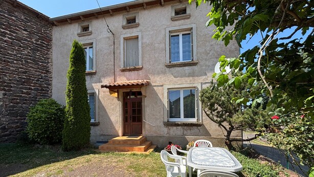 Il s’agit d’un bâtiment à deux étages, doté d’une façade en pierre, d’une porte en bois et d’un petit porche avec des marches. On y trouve une table et des chaises d’extérieur blanches, ainsi qu’un jardin avec différentes plantes et un arbre.