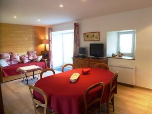 Une pièce chaleureuse avec un plancher en bois, une table de salle à manger rouge et quatre chaises, un canapé agrémenté de coussins à motifs, un bureau avec un ordinateur et un écran, et une fenêtre avec des rideaux.