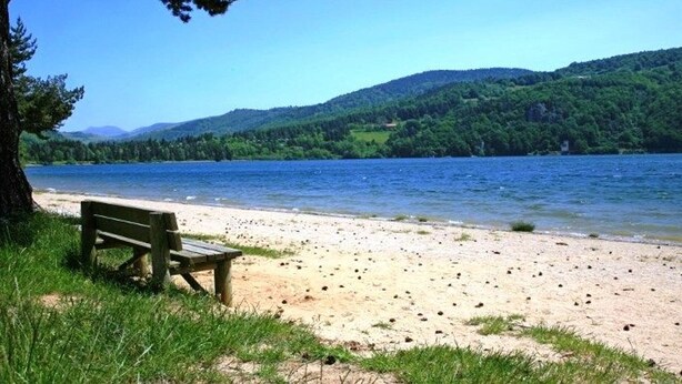 A bench on a sandy beach with a lake and forested hills in the background.