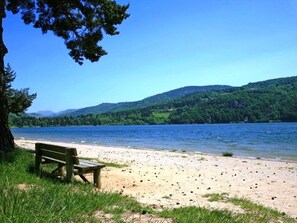 Eine Bank an einem sandigen Strand, im Hintergrund ein See und bewaldete Hügel.